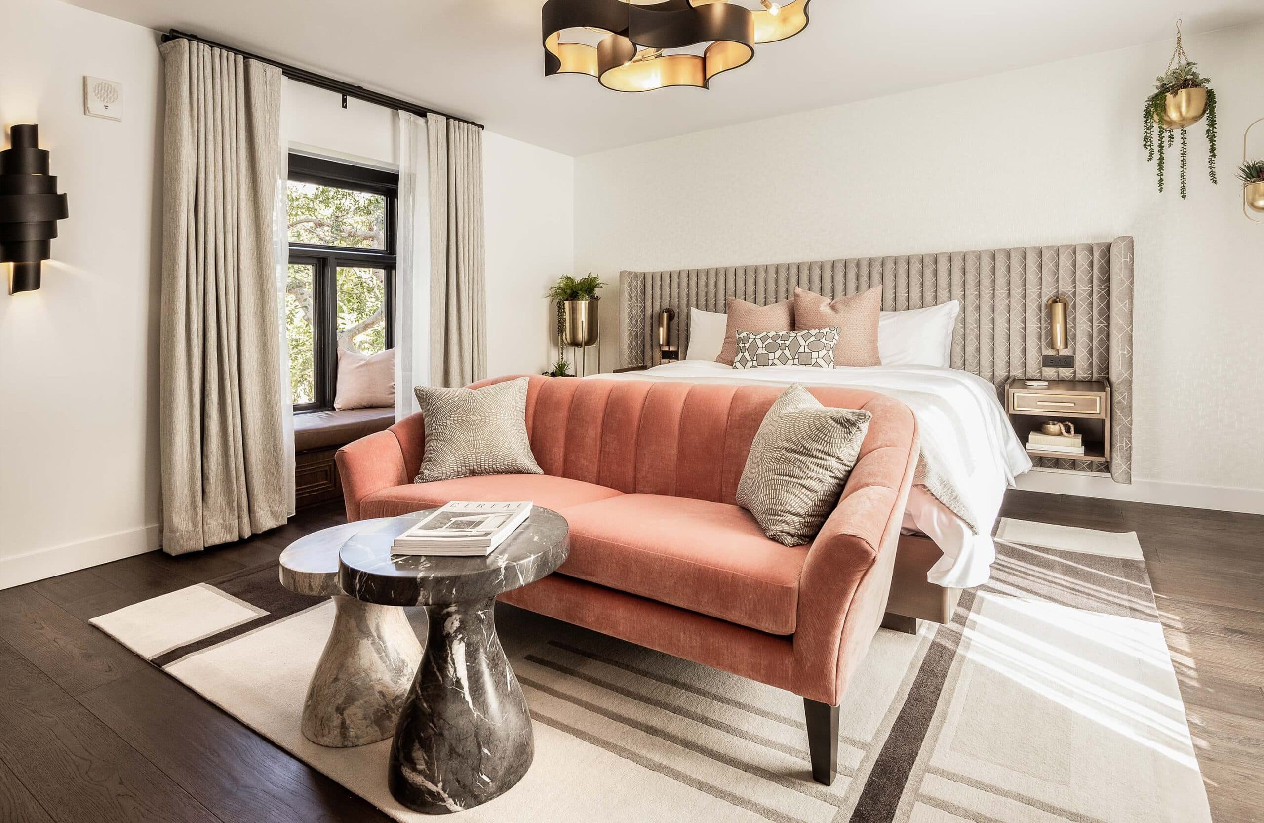Bright modern bedroom with a plush upholstered bed, pink accent sofa, and natural light from large windows.