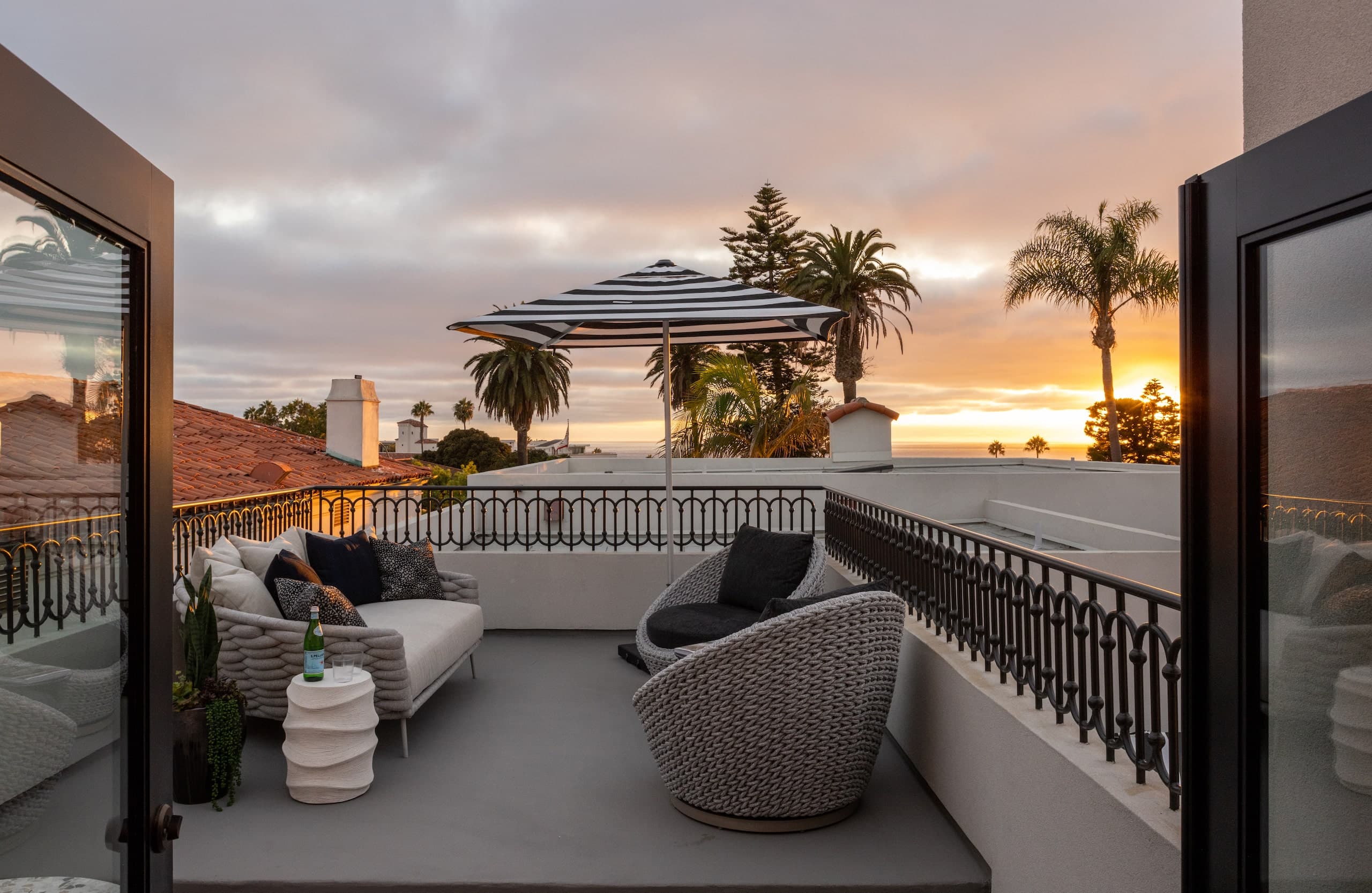 Rooftop terrace with modern outdoor seating, striped umbrella, and palm trees overlooking the ocean at sunset