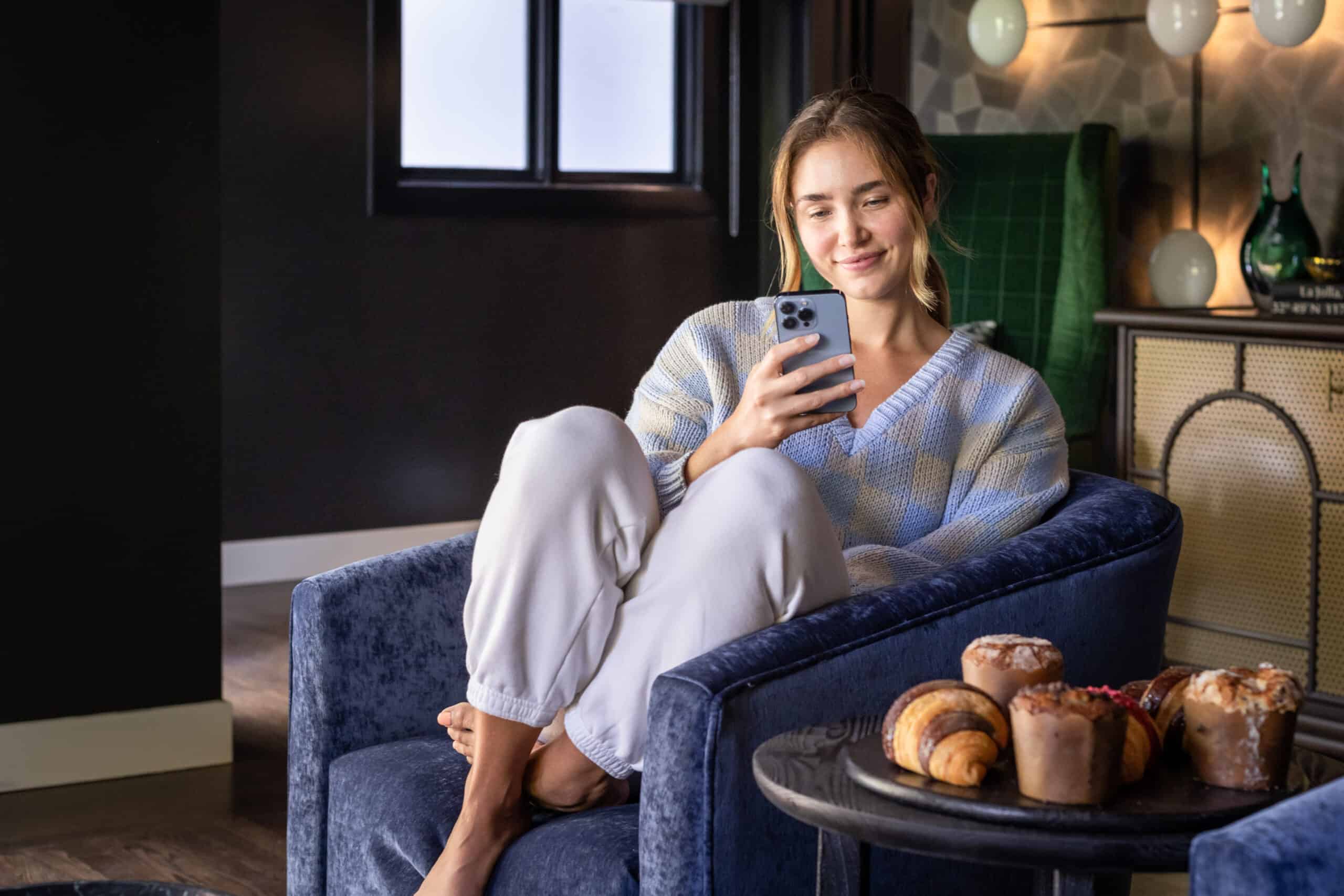 Woman relaxing on a blue armchair while using a smartphone, with fresh pastries nearby in a cozy, modern living space