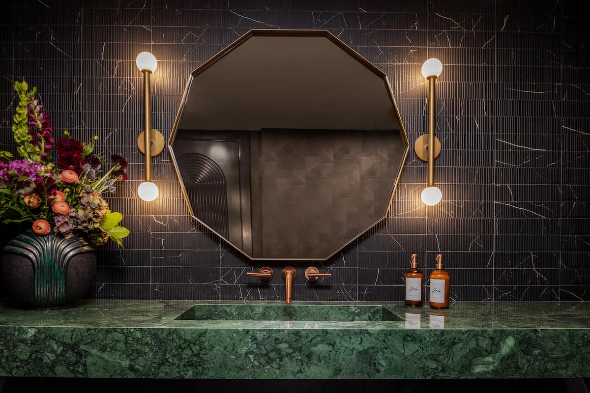 Luxury hotel bathroom vanity with an octagonal mirror, brass sconces, dark tile walls, and a green marble countertop.