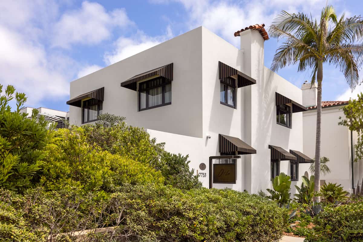 White Mediterranean-style building with black awnings and lush landscaping in a sunny coastal neighborhood