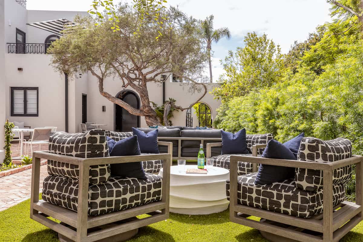 Modern outdoor patio seating area with cushioned sofas, a round fire pit table, and a landscaped courtyard.