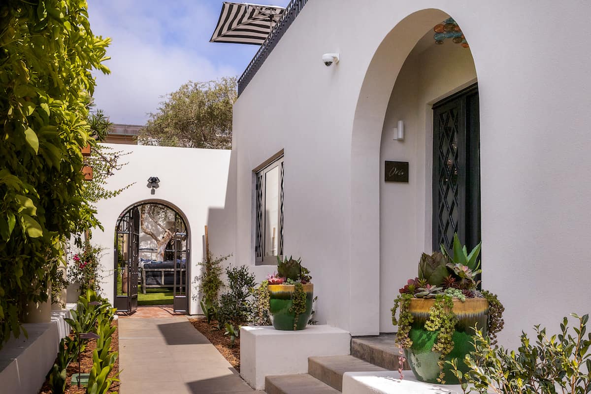 White stucco house exterior with arched doorway, black wrought-iron gate, and potted plants along a sunlit walkway.
