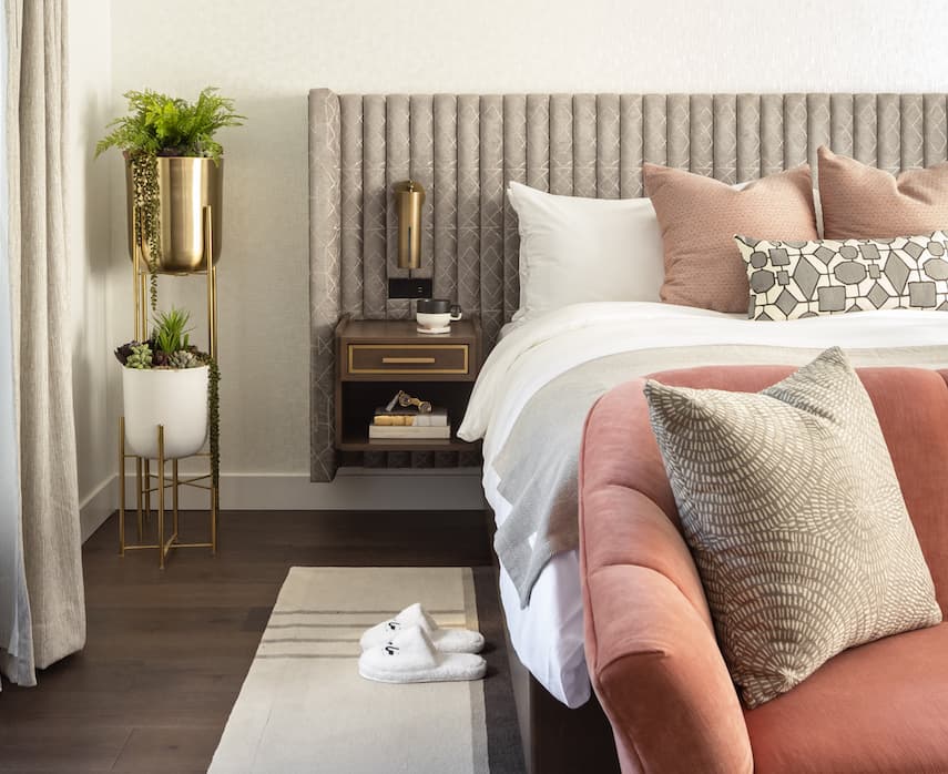 Modern hotel bedroom with upholstered headboard, layered pillows, brass wall lamp, indoor plants, and slippers on a floor rug.