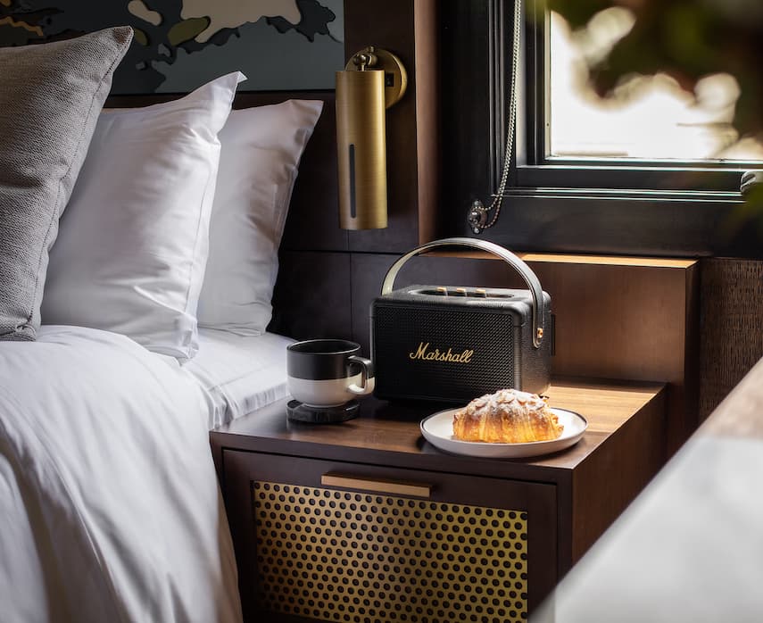 Hotel bedside table with white bedding, a Marshall speaker, coffee cup, and fresh croissant beside a window with natural light.