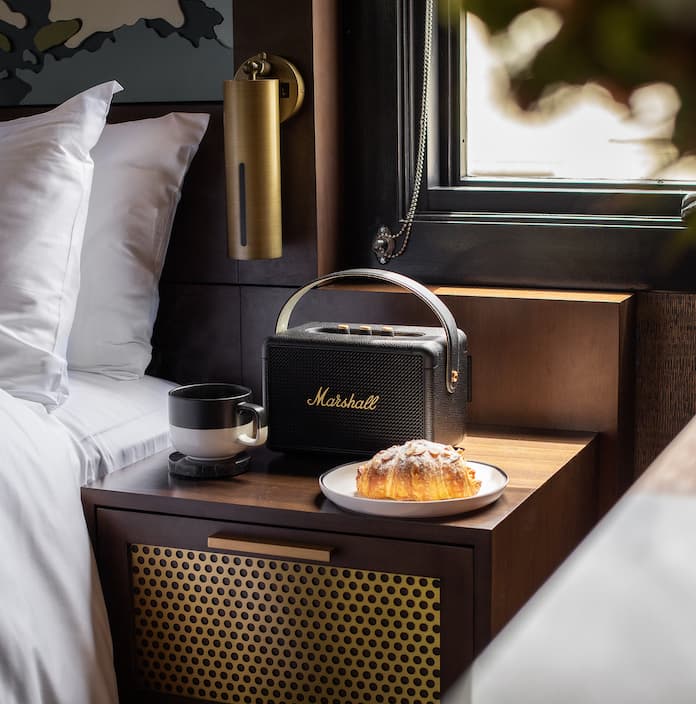 A cup of coffee, a croissant and a bluetooth speaker on a bedside table inside one of the Orli guestrooms