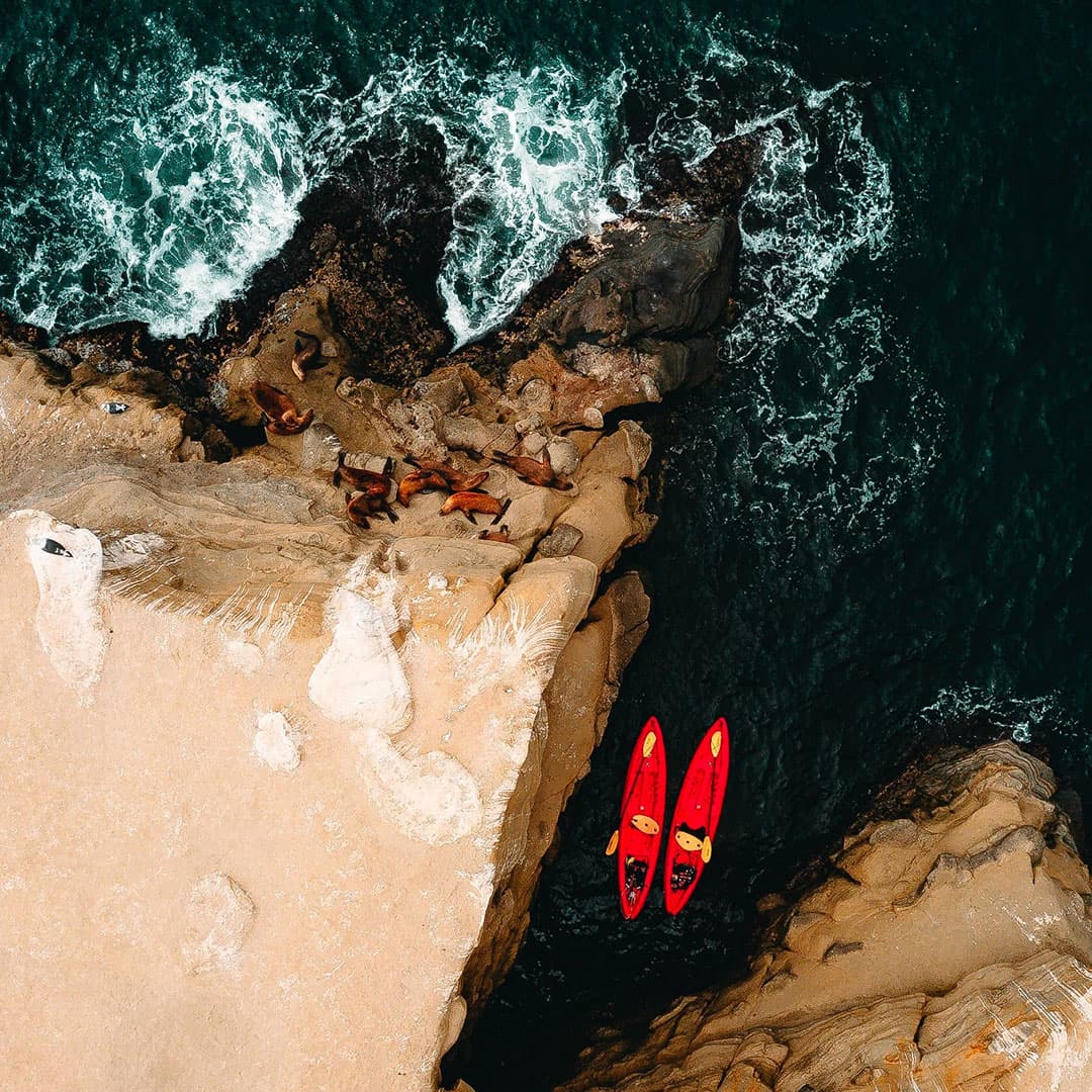 Aerial view of two red kayaks navigating a narrow coastal inlet between rocky cliffs and ocean waves