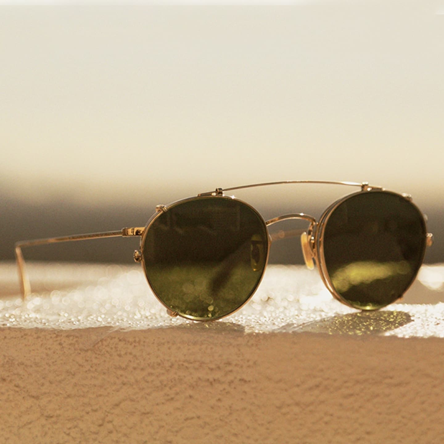 Round gold-framed sunglasses resting on a sandy beach at sunset with warm coastal light