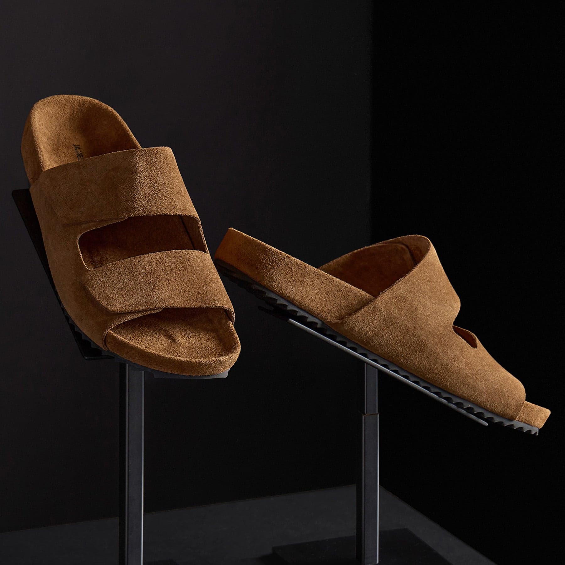 Brown suede slide sandals displayed on minimalist stands against a dark background for premium footwear styling