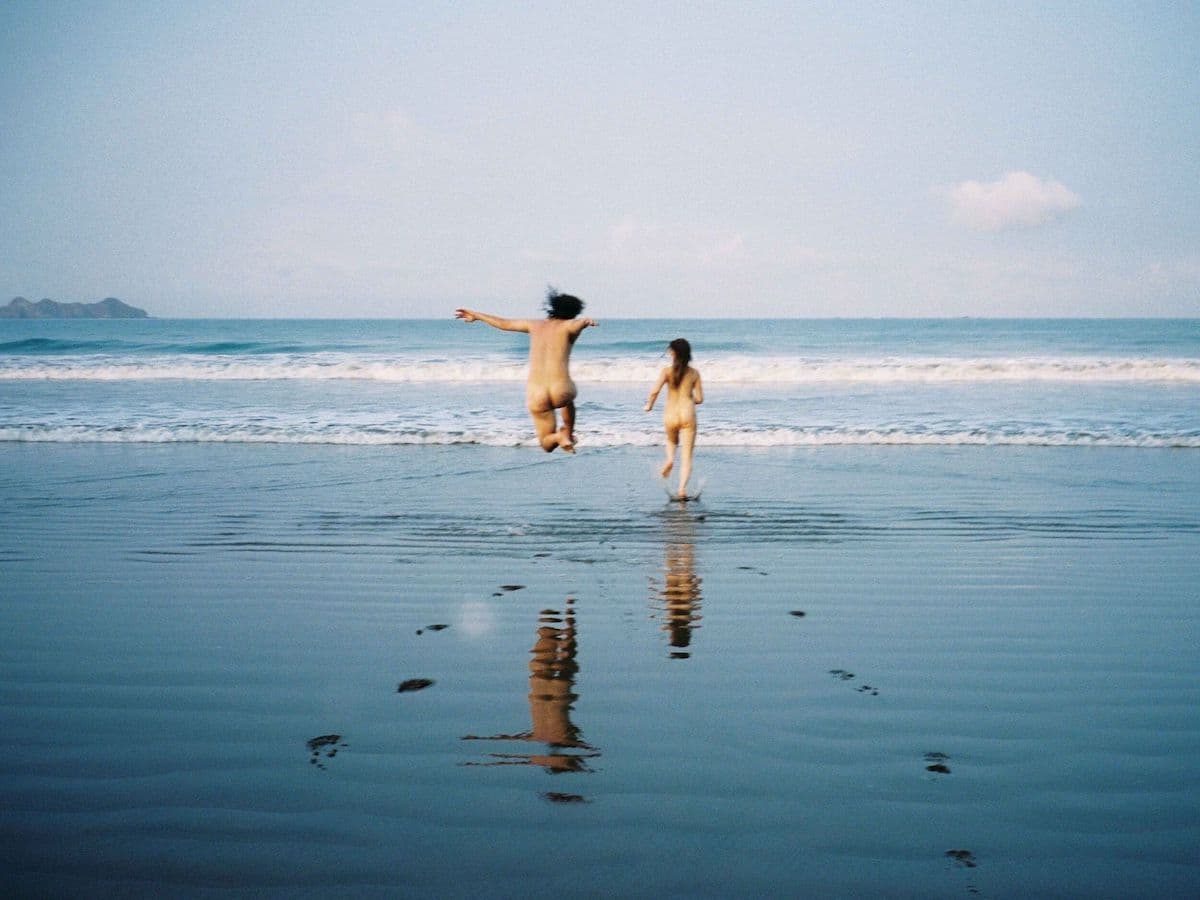 Adult and child holding hands and running toward the ocean along a reflective sandy beach under a clear blue sky