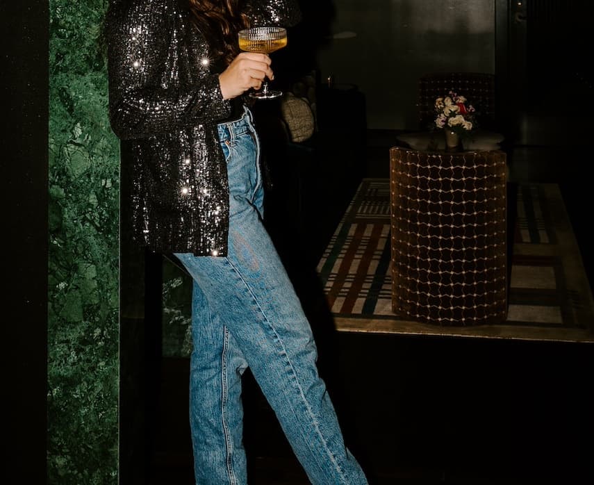 Woman wearing a black sequin top and blue jeans holding a coupe cocktail glass in a dimly lit lounge.