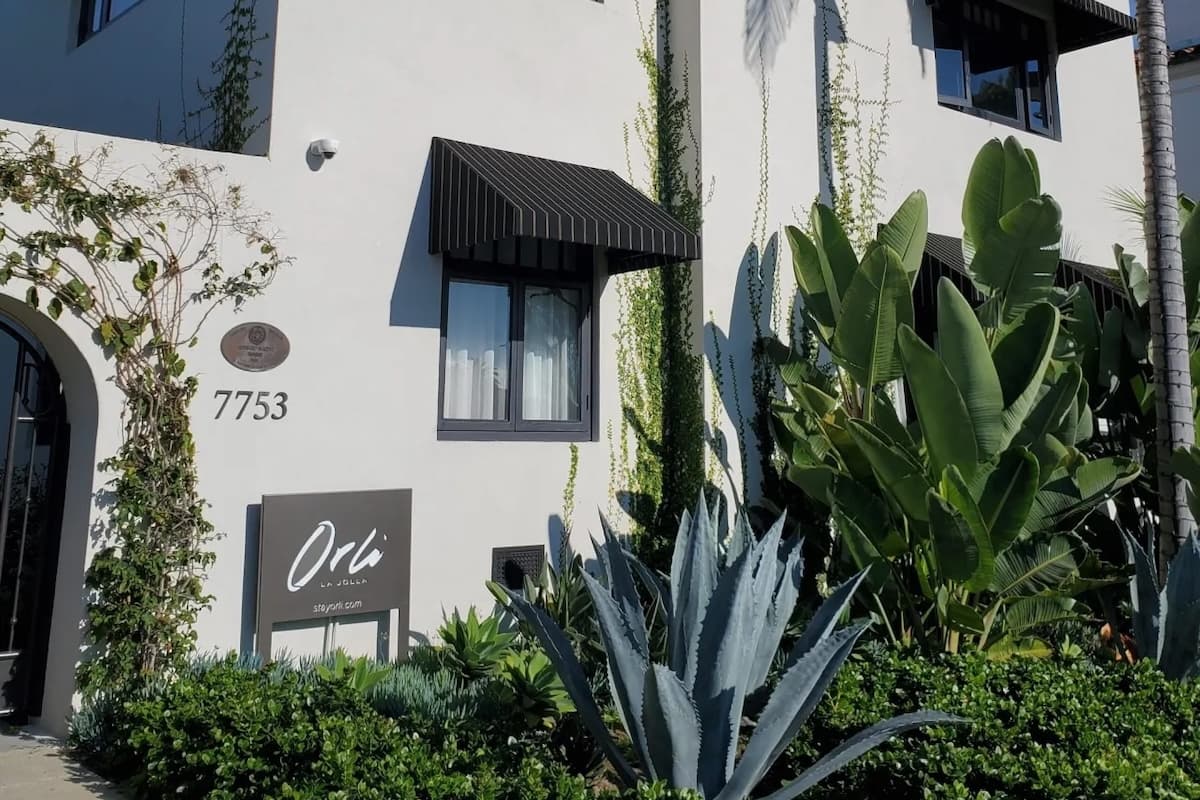 Exterior of the Orli La Jolla hotel featuring white stucco walls, black awnings, and lush tropical landscaping