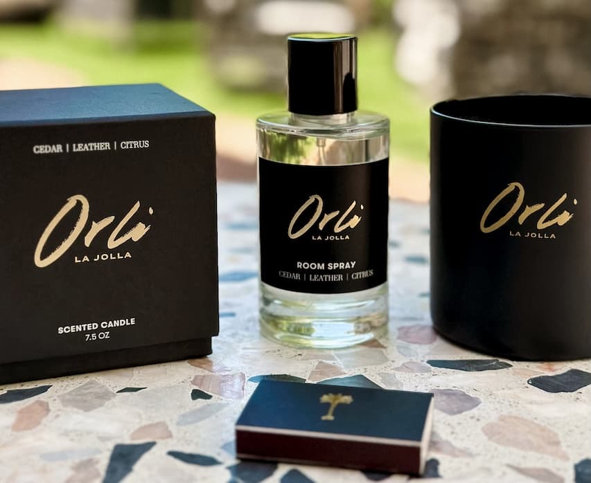 Minimalist product display of Orli La Jolla home fragrance items, including a black boxed scented candle, a cedar leather citrus room spray bottle, a matte black candle jar, and a branded matchbox on a stone tabletop.