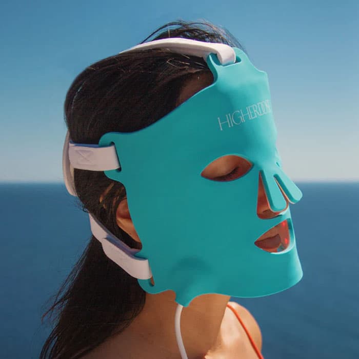Woman wearing a turquoise LED light therapy face mask outdoors with ocean view in the background