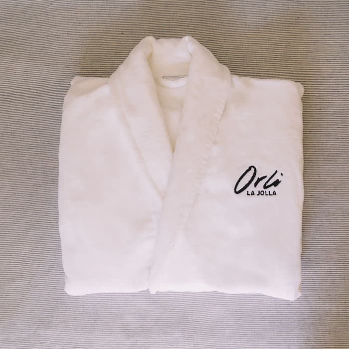 Plush white bathrobe embroidered with Orli La Jolla logo, neatly folded on a textured neutral surface