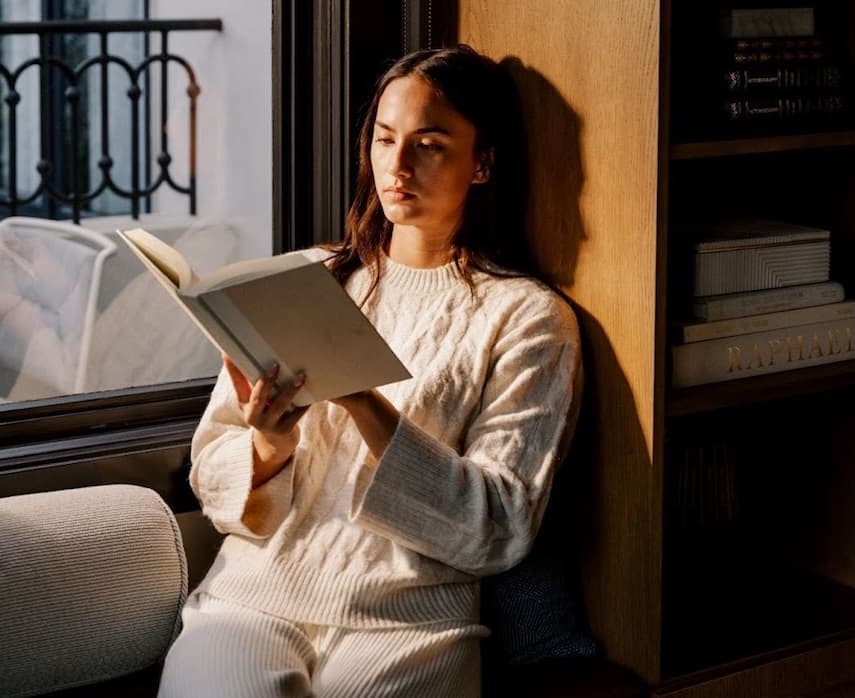 Woman reading a book by a sunlit window in a cozy, modern interior, capturing a calm lifestyle moment focused on relaxation, mindfulness, and comfortable at-home living.