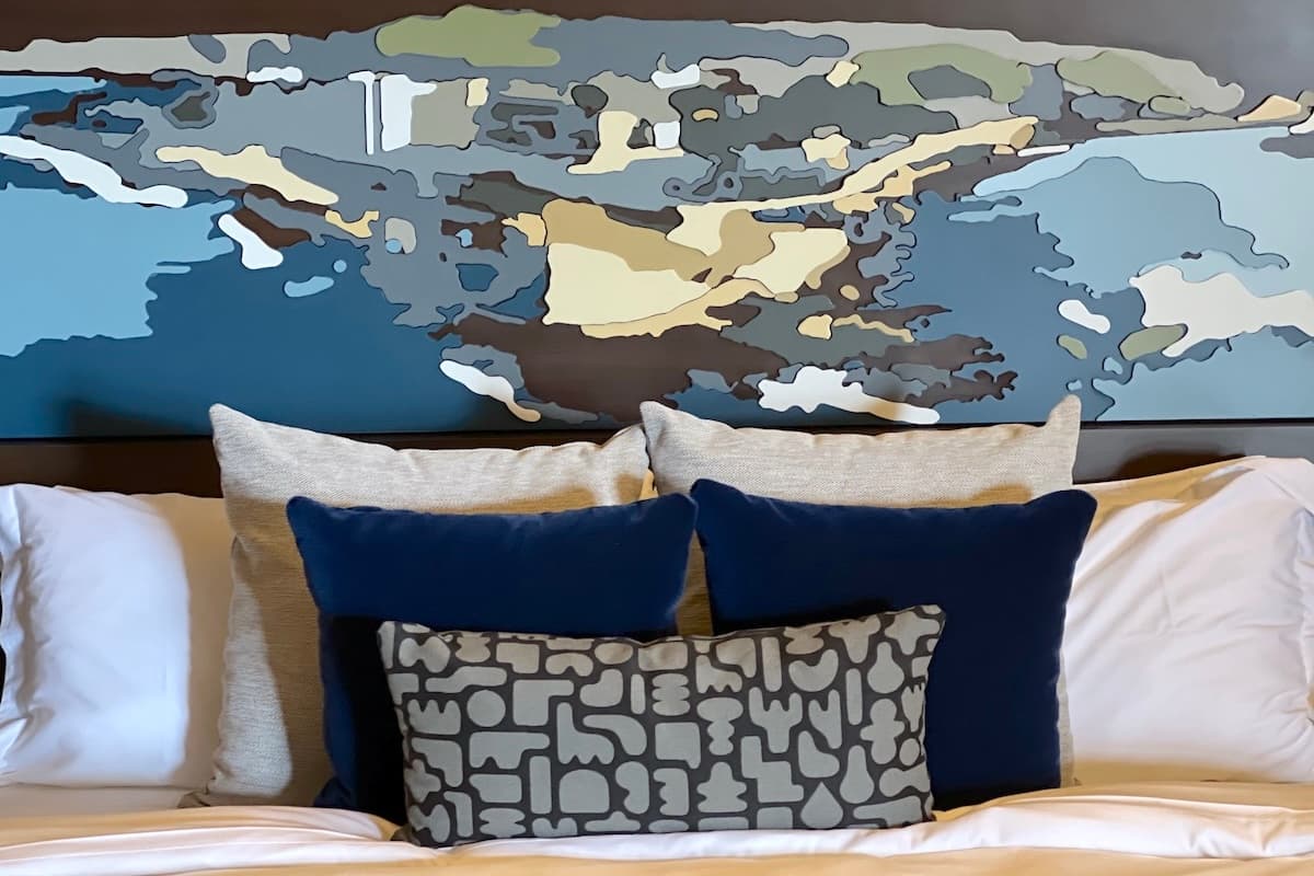 Styled hotel bed with layered white and navy pillows beneath an abstract coastal-inspired wall art headboard