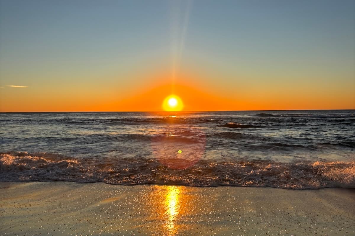 Bright orange sunset over the ocean with waves rolling onto the sandy shore and sunlight reflecting across the water and wet sand.