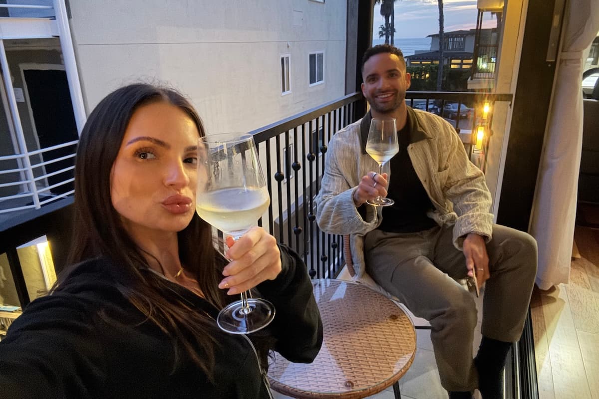 Two people on a balcony at sunset holding glasses of white wine, with palm trees and the ocean visible in the background.