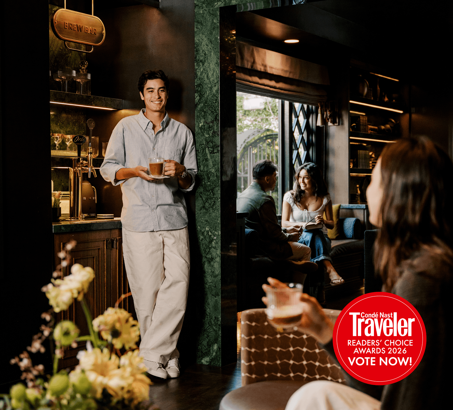 A smiling barista stands at a small brew bar holding a cup of coffee, while a guest sits in the foreground with a drink. In the background, a couple chats on a couch near a window, creating a warm, cozy café atmosphere.