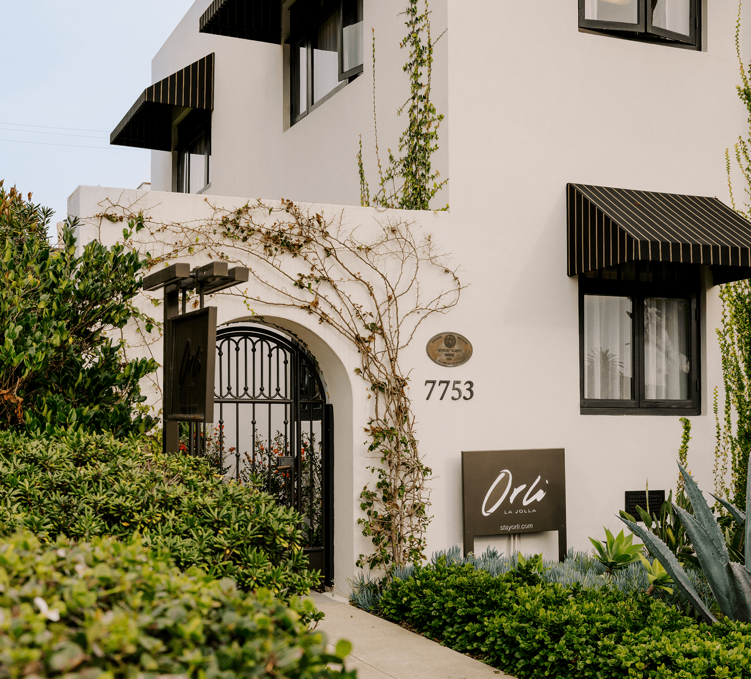Exterior of a white stucco building with black awnings and greenery, featuring a gated entrance and signage reading “Orli” and the address “7753,” surrounded by landscaped plants and vines.
