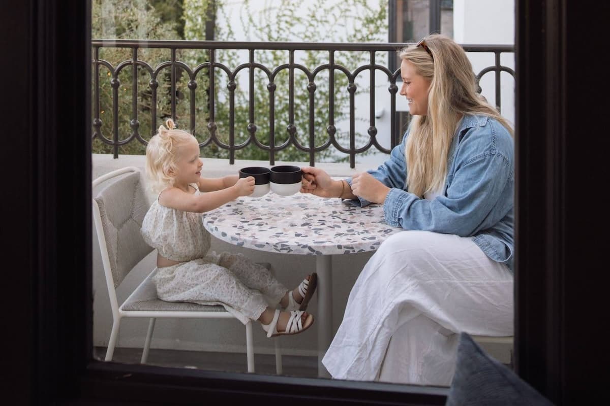 A woman and a young child sitting at a small outdoor table, smiling and clinking cups on a balcony.