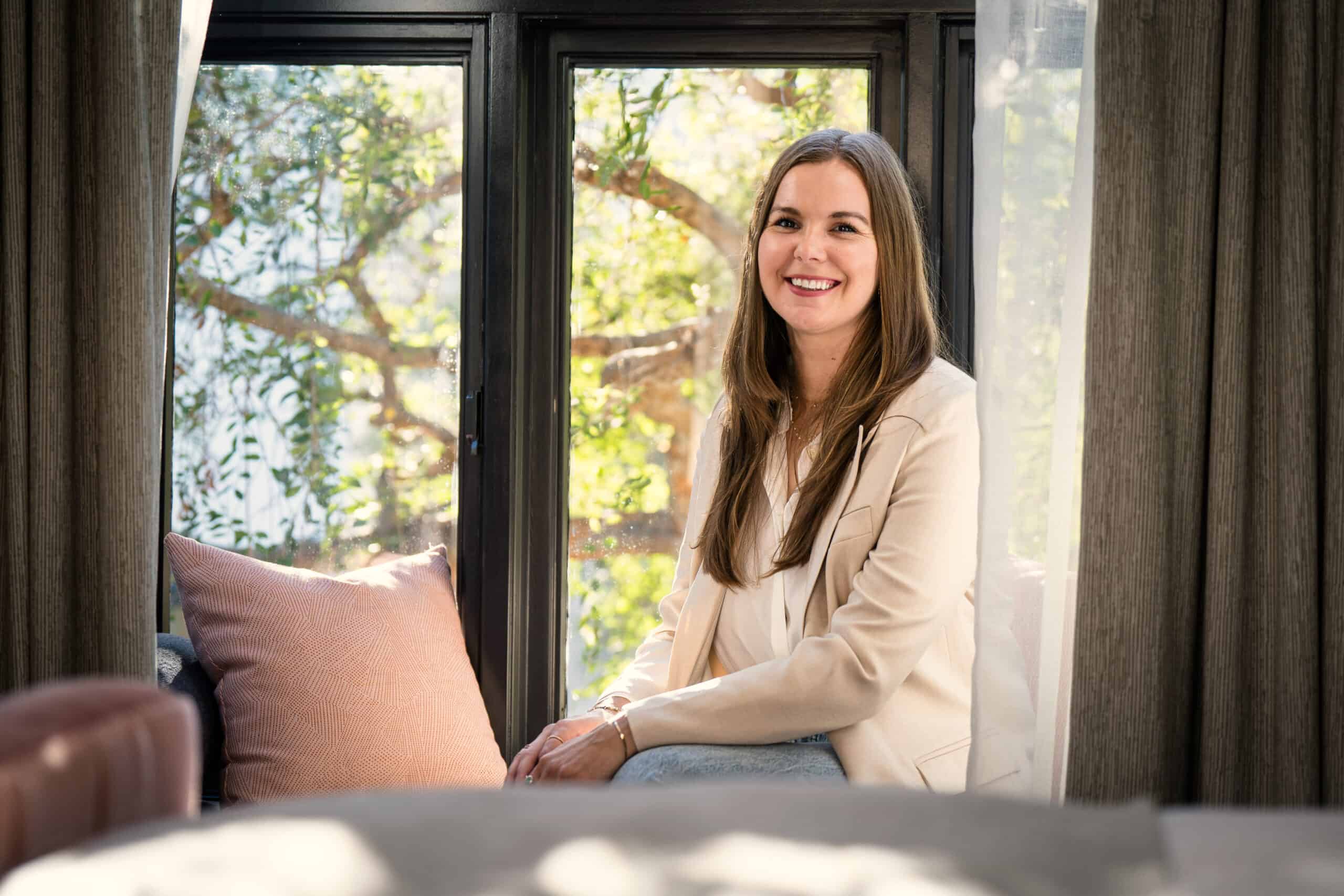 Smiling woman sitting by a window in a softly lit living room with natural light and greenery outside.