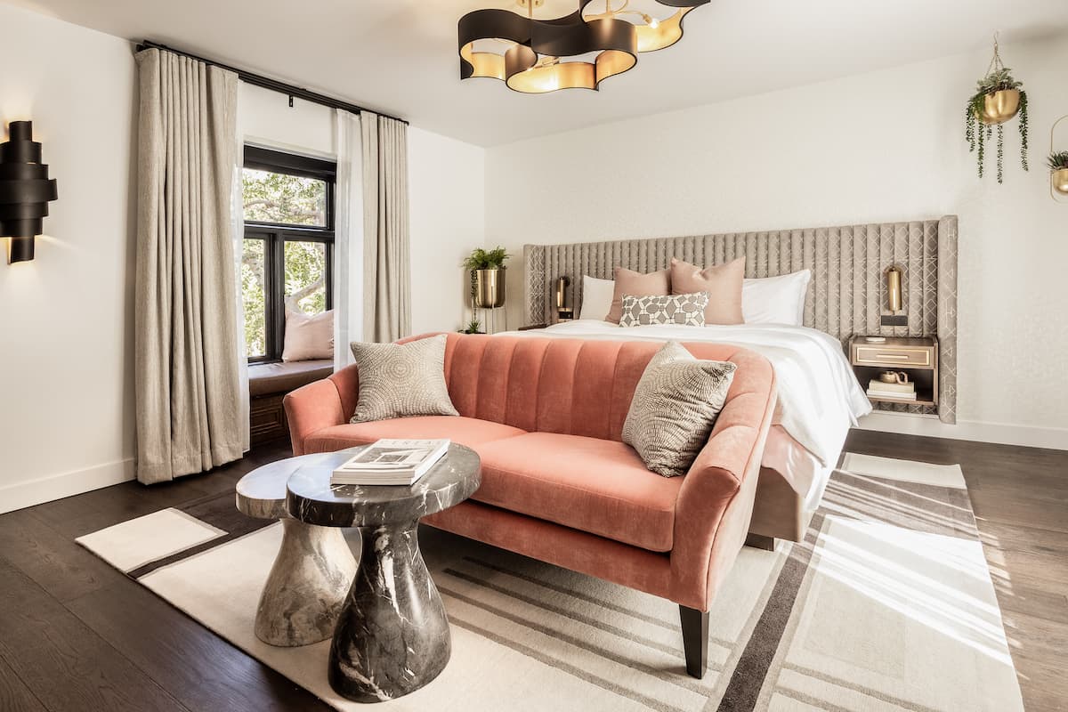Bright luxury hotel suite featuring a plush blush sofa, king-size bed, modern accent tables, and natural light from large windows