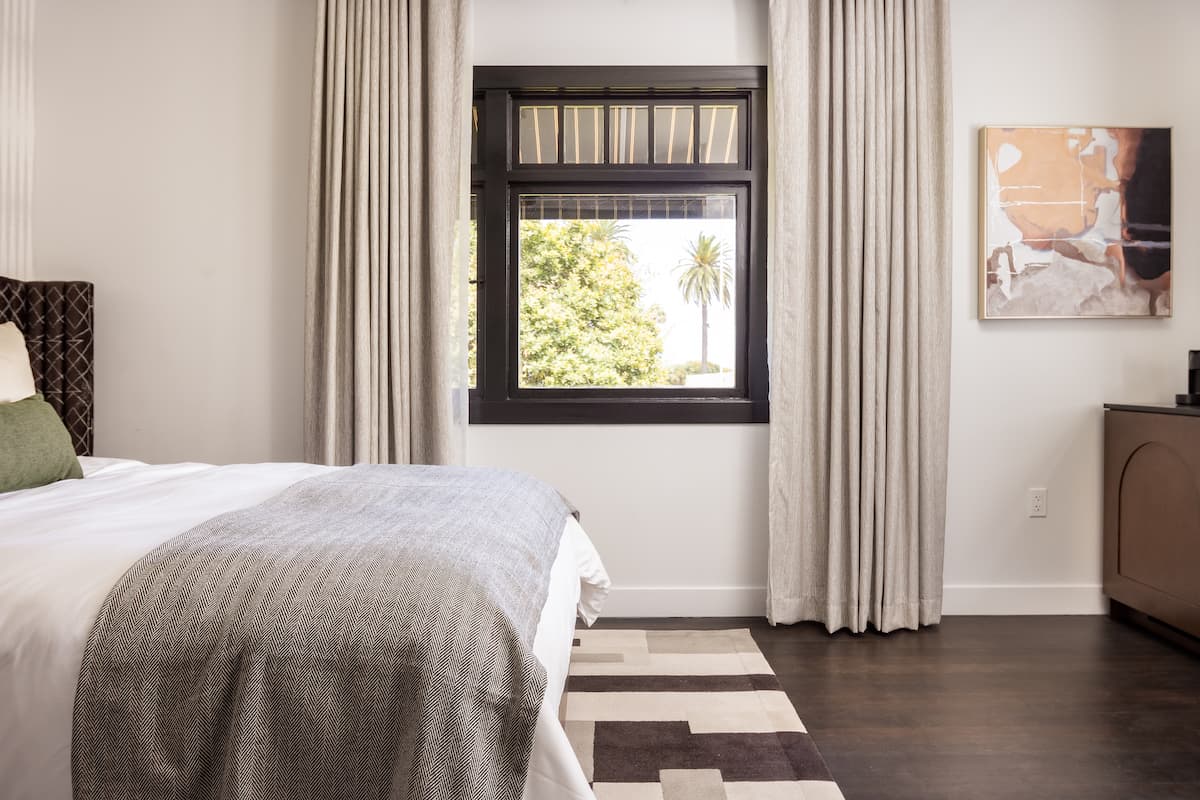 Bright hotel bedroom with neatly made bed, soft neutral bedding, large window with curtains, and modern minimalist décor