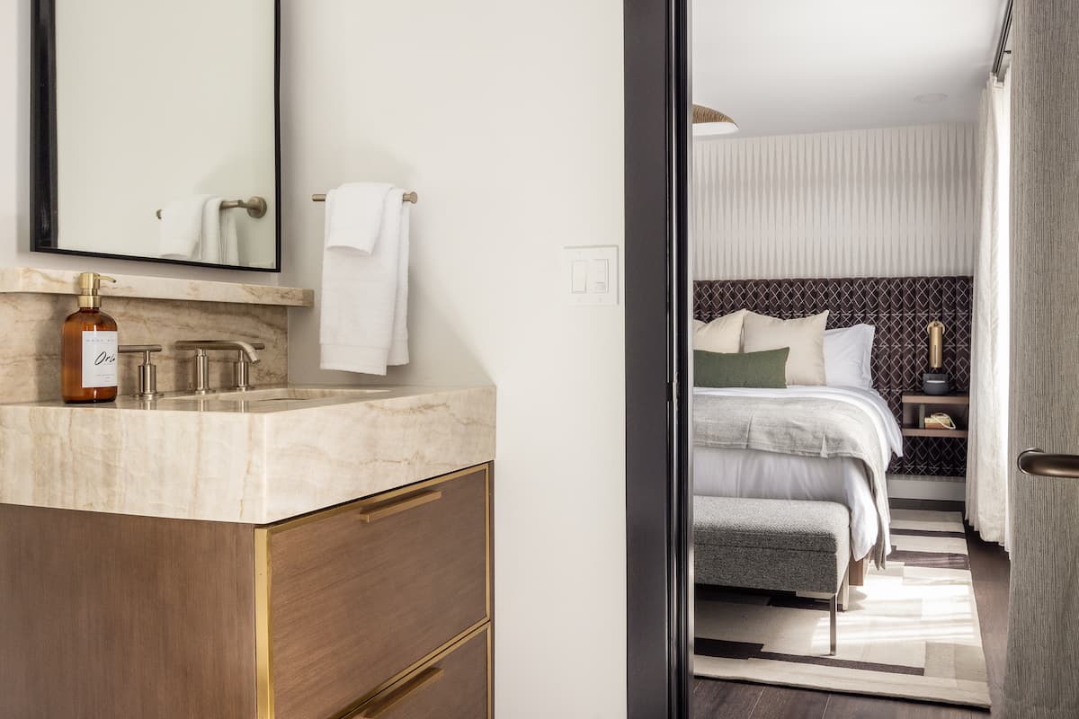 View from a modern hotel bathroom vanity into a stylish bedroom with layered bedding and contemporary décor