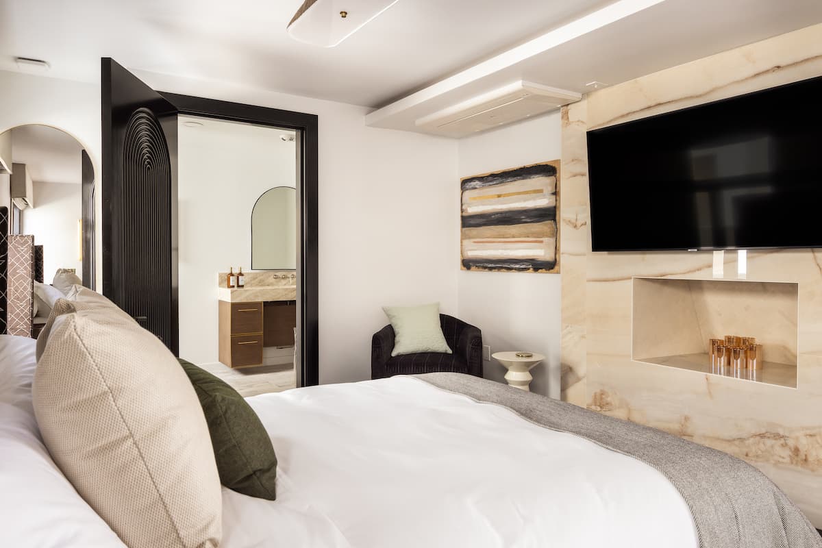 Elegant hotel bedroom with plush white bedding, wall-mounted TV, textured accent wall, and arched doorway to a modern bathroom