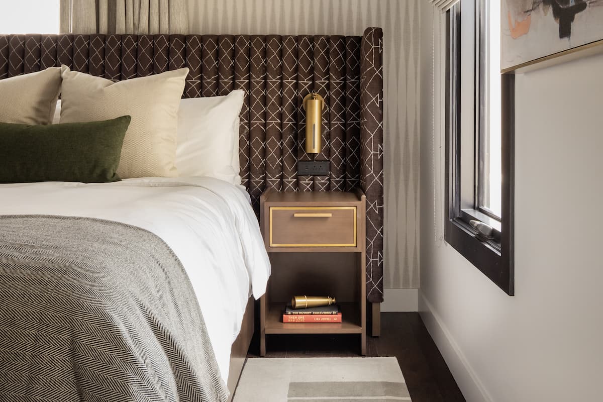 Hotel bedside table with modern lamp, stacked books, and upholstered headboard detail in a refined luxury bedroom