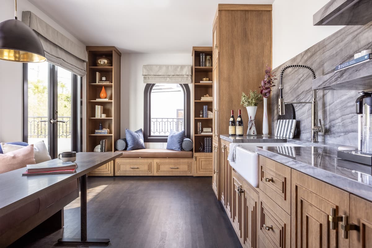 Open-concept luxury suite kitchenette with marble countertops, wooden cabinetry, dining table, and window seating overlooking a balcony