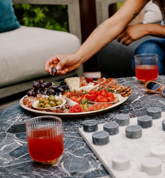 Hand reaching for grapes from a charcuterie board with cheese, cured meats, olives, and fruit on a marble outdoor table, with red drinks and a board game nearby.