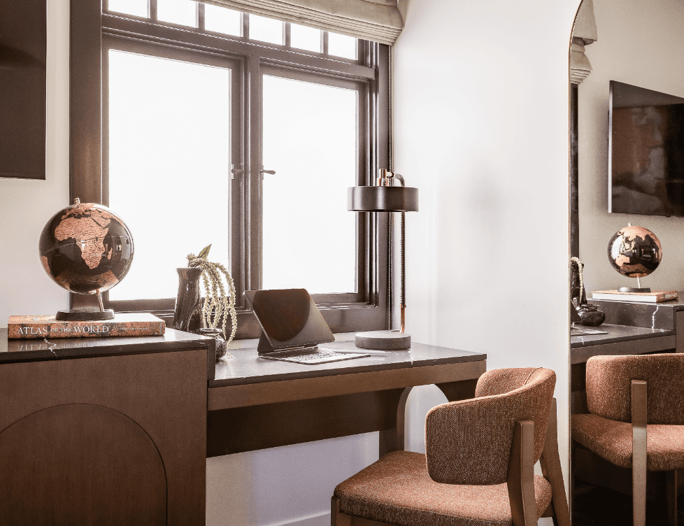 Stylish home office workspace with desk, laptop, globe decor, upholstered chair, and large window with natural light.