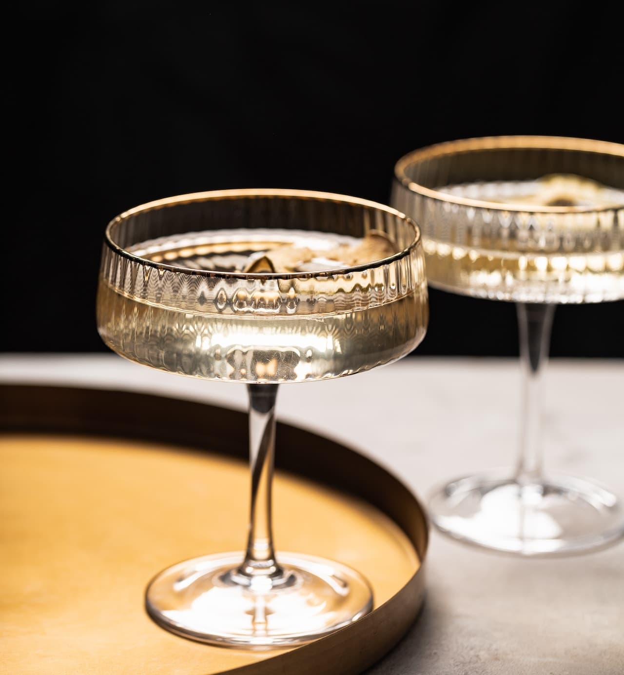 Two coupe cocktail glasses with a pale drink on a gold tray against a dark background.