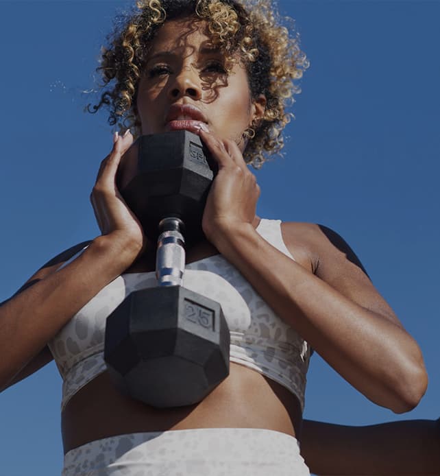 Athletic woman lifting a dumbbell outdoors against a clear blue sky, wearing a sports bra and focused expression.