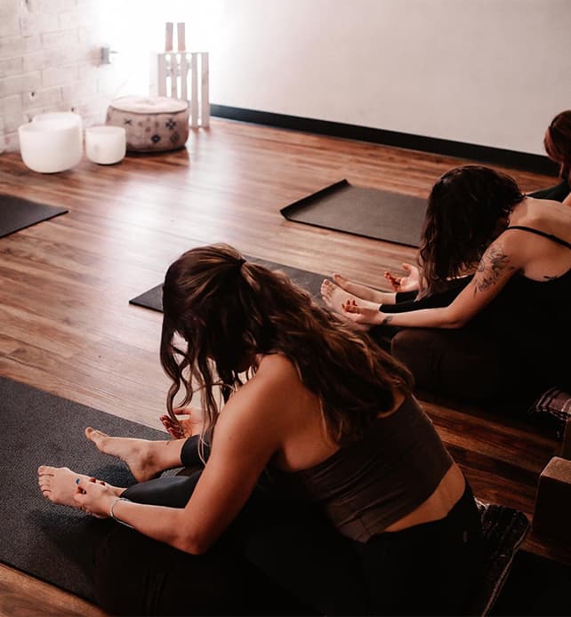 Small group yoga class practicing seated stretches on mats in a softly lit studio with wooden floors.