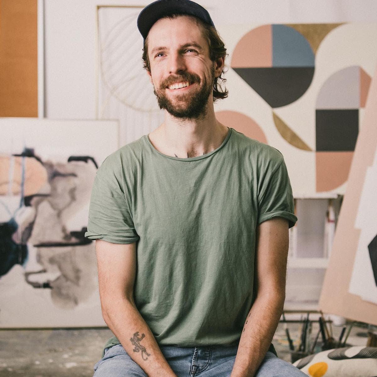 Smiling man wearing a green t-shirt and cap, seated in an art studio surrounded by abstract artwork