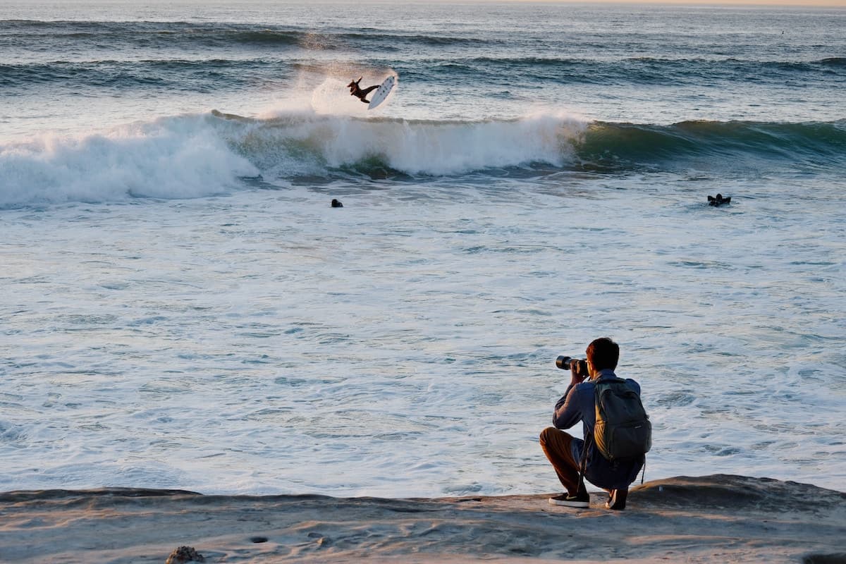 Photographer kneeling on coastal rocks capturing a surfer launching above a breaking ocean wave.