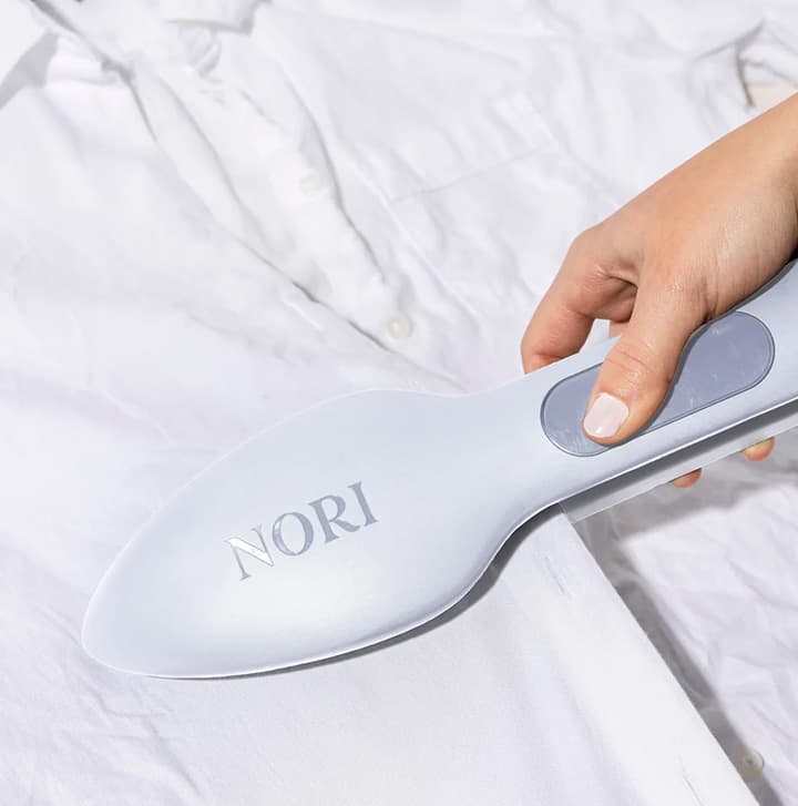 Handheld Nori fabric steamer smoothing wrinkles on a white shirt, showcasing a modern garment care tool
