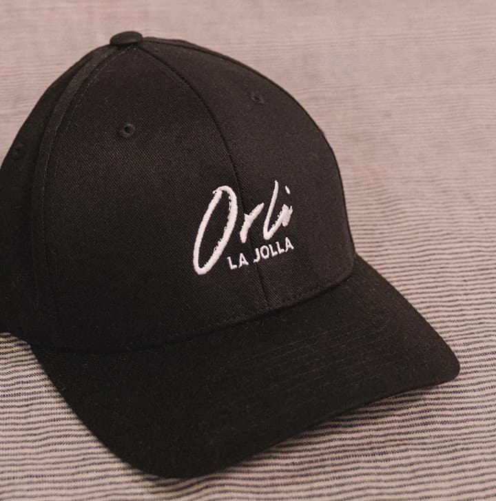 Black Orli La Jolla baseball cap with white embroidered logo, displayed on a neutral textured surface