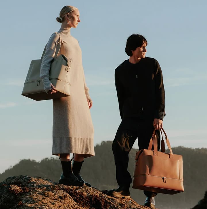 Two people standing outdoors on rocky terrain holding structured leather tote bags, styled for a modern lifestyle fashion shoot