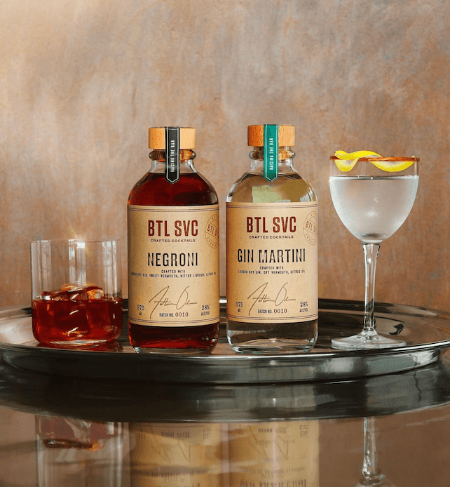 Bottled Negroni and gin martini cocktails displayed on a reflective tray beside a coupe glass with a lemon twist.