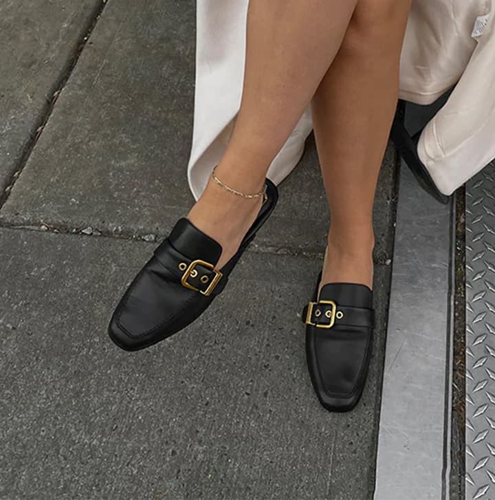 Woman wearing black leather loafers with gold buckle detail, styled with a light dress on a city sidewalk