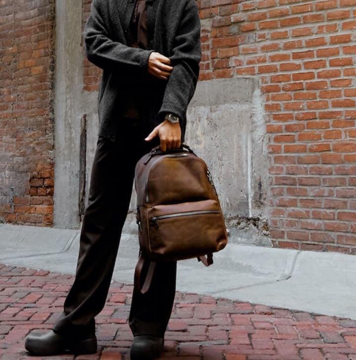 Person holding a brown leather backpack while standing on a brick sidewalk against an urban brick wall backdrop