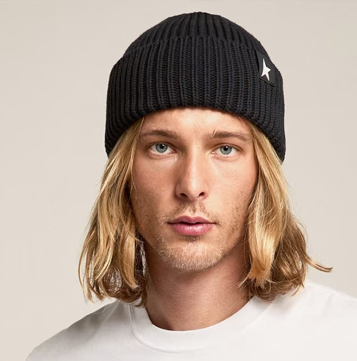 Man with long blond hair wearing a black knit beanie, photographed against a neutral studio background