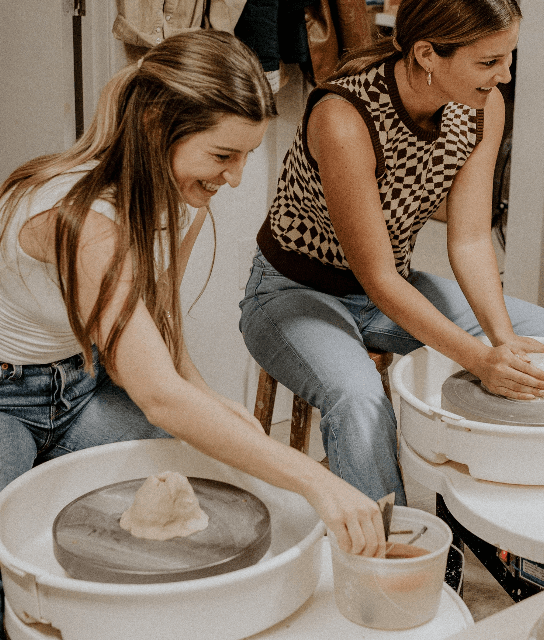 Two women smiling and laughing while working at pottery wheels indoors, shaping clay with their hands during a casual, creative workshop.