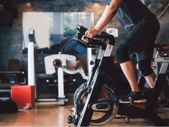 Person pedaling a stationary exercise bike in a modern gym, with fitness equipment and mirrors visible in the background, capturing an indoor cardio workout scene.
