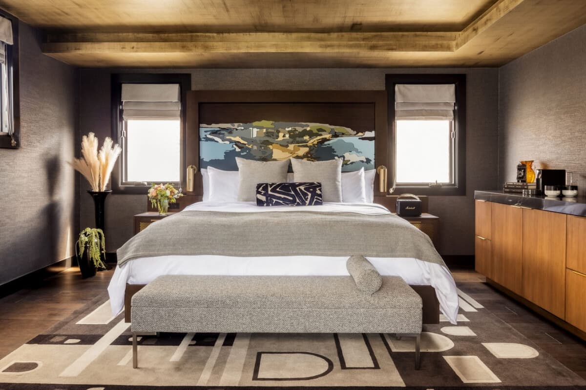 Modern luxury bedroom with a king-size bed, layered neutral bedding, abstract headboard art, wood-paneled ceiling, matching windows, and minimalist furnishings in a warm, upscale hotel-style interior.